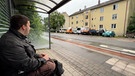 Ein Mann sitzt auf einer Bank in einer Bus-Haltestelle. | Bild: Bayerischer Rundfunk 2024