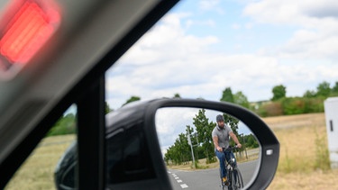 Auf einem Testgelände fährt ein Fahrradfahrer neben einem Auto mit einem Abbiegeassistenten vom Technologieunternehmen Continental her. Das Fahrzeug erkennt den Mann, ein rotes Licht leuchtet und das Auto bremst automatisch.  | Bild: picture alliance/Christophe Gateau/dpa