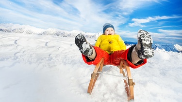 Ein Kind fährt mit ausgestreckten Beinen mit einem Schlitten auf den Betrachter zu. | Bild: colourbox.com