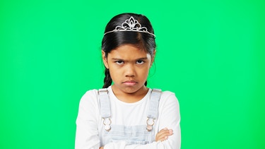 Ein Mädchen mit schwarzen Haaren und kleiner Prinzessinenkrone steht mit verschränkten Armen vor grünem Hintergrund und schaut trotzig in die Kamera. | Bild: colourbox.com