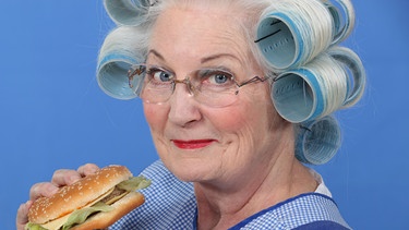 Eine Seniorin mit Lockenwicklern im Haar und einem Burger in der Hand | Bild: colourbox.com