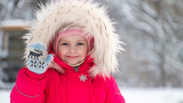 Ein Mädchen mit dickem Winteranorak und Fellkapuze winkt mit roten Backen in die Kamera. | Bild: colourbox.com