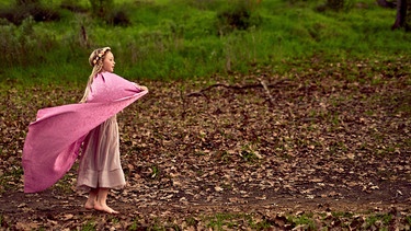 Ein Mädchen steht mit einem rosanen Umgang und einem Blütenkranz auf einer Lichtung im Wald. | Bild: colourbox.com