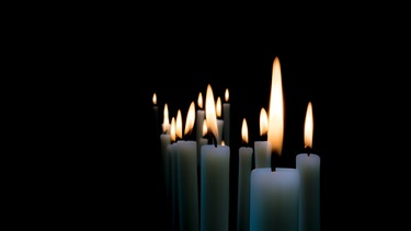 Brennende weiße Kerzen vor schwarzem Hintergrund. | Bild: stock.adobe.com/blickpixel