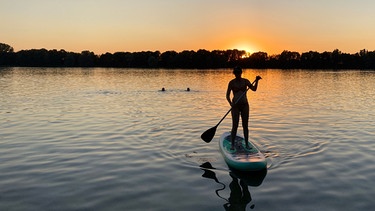 Eine junge Frau auf einem Stand-Up Paddle bei Sonnenuntergang. | Bild: BR/Annette Goossens