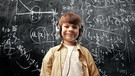 Ein Junge mit großen Kopfhörern auf dem Kopf steht vor einer Schiefertafel auf der mathematische Formeln stehen. | Bild: stock.adobe.com/olly