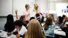Schülerinnen und Schüler sitzen in einem Klassenzimmer. An der Tafel steht eine Lehrerin.  | Bild: stock.adobe.com/rawpixel.com