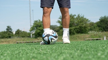 Eine Person, bis zu den Knien sichtbar, steht auf Kunstrasen. Vor der Person liegt ein Fußball auf dem Rasen. | Bild: BR