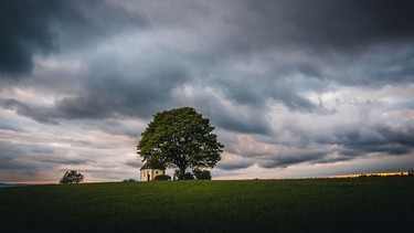 Dramatische Wolken in der Abenddämmerung über einer Wiese mit Kapelle und Baum. | Bild: stock.adobe.com/Thilo Wagner