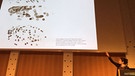 29.12.2018, Sachsen, Leipzig: Die Berliner Künstlerin Liat Grayver stellt auf dem 35. Chaos Communication Congress (35c3) Bilder vor, die der Roboter«e-David» gemalt hat. Die Maschine wurde von Wissenschaftlern der Universität Konstanz entwickelt.  | Bild:  Peter Zschunke/dpa