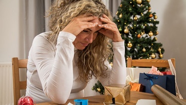 Eine Frau schaut gestresst auf Geschenke und Verpackungen. Im Hintergrund steht ein Weihnachtsbaum. | Bild: picture alliance / dpa-tmn | Christin Klose