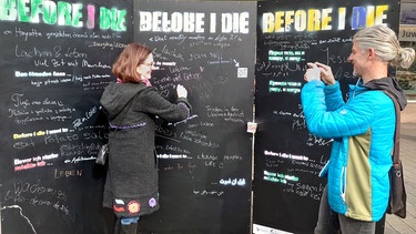 Susanne Bammessel und Thomas Amberg stehen vor drei schwarzen Tafeln der Kunstaktion "Before I die". | Bild: BR24/Ulrike Nikola