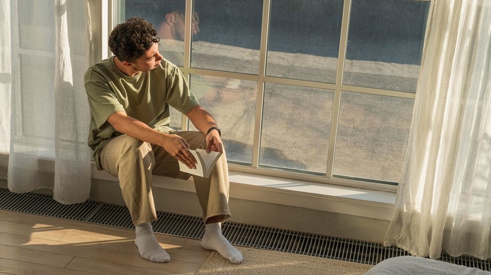 Symbolbild Einsamkeit junge Menschen. Junger Mann sitzt mit Buch am Fenster. | Bild: picture alliance / Westend61 | Vasily Pindyurin