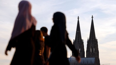 Frauen mit Kopftuch am Kölner Rheinboulevard, im Hintergrund ragt die Doppelspitze des Kölner Doms in den Himmel.  | Bild: picture alliance / Panama Pictures | Christoph Hardt