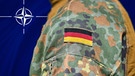 Ein Soldat in deutscher Tarnuniform mit aufgenähter Deutschlandflagge steht vor einer Nato-Flagge. | Bild: picture alliance / CHROMORANGE | Michael Bihlmayer