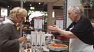 Gesprächssituation zwischen einer Kundin und einer Verkäuferin von Kochgeschirr. Sie präsentiert der Kundin Speisen, die sie frisch gebacken hat.  | Bild: Lukas Graw / BR