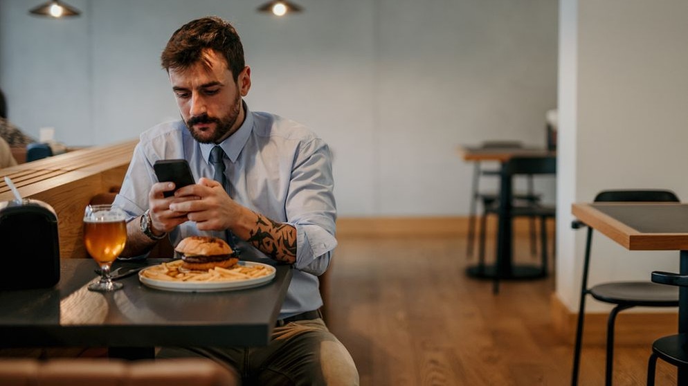 Ein Mann sitzt in einem Restaurant bei einem Burger und einem Bier, hält ein Smartphone in der Hand und tippt darauf eine Nachricht. | Bild: stock.adobe.com/La Famiglia