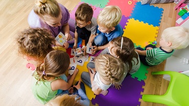 Eine Kindergartengruppe sitzt zusammen mit der Erzieherin auf dem Boden und lernt die Buchstaben. | Bild: stock.adobe.com