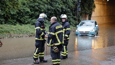 Drei Feuerwehrleute stehen neben einer überfluteten Unterführung, im Hintergrund ein Auto im Wasser. | Bild: NEWS5/David Oßwald