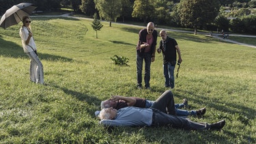 Vorne: Udo Wachtveitl (Rolle: Kriminalhauptkommissar Franz Leitmayr) und Miroslav Nemec (Rolle: Kriminalhauptkommissar Ivo Batic) liegen im Gras. Stehend: Regisseur Andreas Kleinert (Mitte) und Kameramann Johann Feindt überlegen, wie sie diesen Moment am besten mit der Kamera einfangen können. Links: Ein Crewmitglied sieht ihnen dabei zu. | Bild: BR/Wiedemann & Berg Television GmbH & Co. KG/Hendrik Heiden