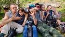 Von links: Die Schüler und Schülerinnen Luca, Theo, Maxi, Max, Sophia, Anabelle und Marlies posieren auf dem Schulgelände beim Wanderschuhe anprobieren. Das „W“ steht für „Wanderstiefel“! Und diese müssen gepflegt werden. Mit einer ordentlichen Wachsschicht halten die Schuhe länger dicht. | Bild: BR/Bewegte Zeiten Filmproduktion GmbH/Megan Ehrmann