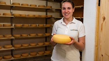 Magdalena Kaiser steht in ihrem Reiferaum und hält ein Laib selbstgemachten Käse in der Hand. | Bild: BR/megaherz gmbh/Moritz Sonntag