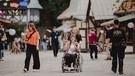 Mehrere Menschen auf dem PULS Open Air-Gelände. In der Mitte des Fotos ist eine Frau im Rollstuhl zu sehen, die Essen in beiden Händen hochhält und lächelt. Impressionen vom PULS Open Air 2024 auf Schloss Kaltenberg. | Bild: BR/Johanna Schlüter