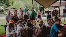 Mehrere Festivalbesucher:innen sitzen unter einem Zelt der News-WG-Minispiele. | Bild: BR/Julia Knoblauch