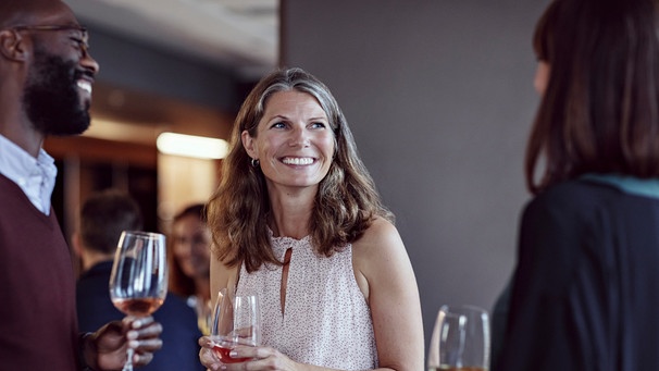 Eine Frau steht in einer Gruppe mit einem Glas Wein in der Hand und lächelt. | Bild: mauritius images / Maskot