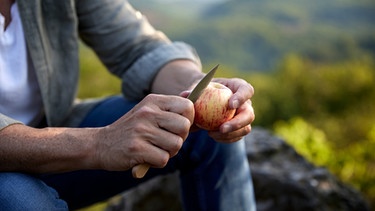 Hände eines Mannes schneiden unter freiem Himmel einen Apfel auf | Bild: mauritius images / Westend61 / Jo Kirchherr