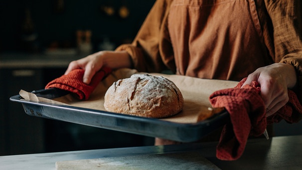 Auf einem Blech liegt Backpapier und ein frisch gebackenes Brot. Eine Frau trägt es mit zwei roten Geschirrtüchern zur Arbeitsfläche in der Küche.  | Bild: mauritius images / Westend61 / Vira Simon