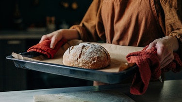 Auf einem Blech liegt Backpapier und ein frisch gebackenes Brot. Eine Frau trägt es mit zwei roten Geschirrtüchern zur Arbeitsfläche in der Küche.  | Bild: mauritius images / Westend61 / Vira Simon