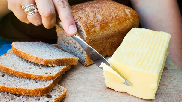 Die Hand einer Frau schneidet ein Stück Butter ab, daneben frische Brotscheiben | Bild: mauritius images / Mint Images Limited / Alamy / Alamy Stock Fotos