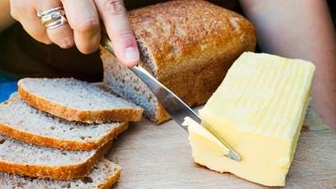 Die Hand einer Frau schneidet ein Stück Butter ab, daneben frische Brotscheiben | Bild: mauritius images / Mint Images Limited / Alamy / Alamy Stock Fotos