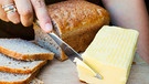 Die Hand einer Frau schneidet ein Stück Butter ab, daneben frische Brotscheiben | Bild: mauritius images / Mint Images Limited / Alamy / Alamy Stock Fotos