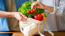 Hand holt aus einer Einkaufstasche mit Gemüse eine Rispe Tomaten heraus | Bild: mauritius images / Prostock-studio / Alamy / Alamy Stock Photos