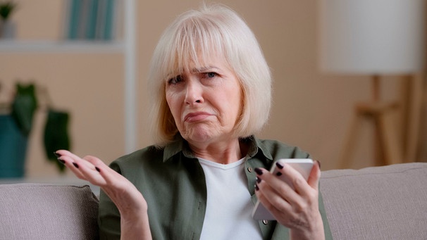 Frau hält das Handy in der Hand und blickt fragend in die Kamera | Bild: mauritius images / Yuliia Kaveshnikova / Alamy / Alamy Stock Photos