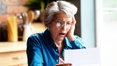 Eine Frau sitzt in einer Wohnküche und sieht entsetzt auf das Schreiben in ihrer Hand. | Bild: mauritius images / Aleksandr Davydov / Alamy / Alamy Stock Photos