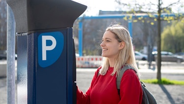 Eine Frau steht an einem Parkscheinautomaten und löst ein Ticket. | Bild: mauritius images / Westend61 / Bernd Friedel