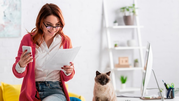 Eine Frau hat ihr Handy und ein Stück Papier in der Hand und freut sich | Bild: mauritius images / LightField Studios Inc. / Alamy / Alamy Stock Photos