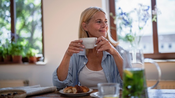 Eine blonde Frau blickt lächelnd zur Seite. Sie sitzt am Frühstückstisch und hält eine Tasse mit beiden Händen fest. | Bild: mauritius images / Westend61 / Richárd Bellevue