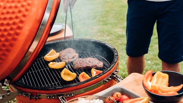 Ein Mann steht vor einem Grill und will mit einer Zange das darauf liegende Fleisch wenden | Bild: mauritius images / Studio Lucky / Alamy / Alamy Stock Photos