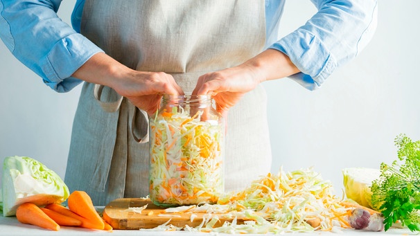 Frau füllt kleingeschnittenen Weißkohl und geraspelte Möhren in ein Einmachglas, um das Ganze zu fermentieren. | Bild: mauritius images / NATALIA KHIMICH / Alamy / Alamy Stock Fotos