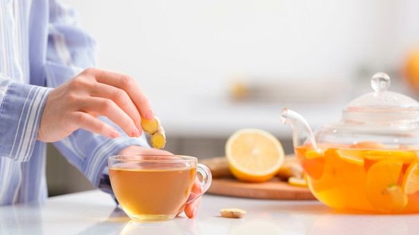 Die Hand einer Frau hält eine Scheibe Ingwer über eine Tasse Ingwertee, im Hintergrund die Teekanne und eine halbe Zitrone auf einem Schneidebrett | Bild: mauritius Bilder / Pixel-shot / Alamy / Alamy Stock Fotos