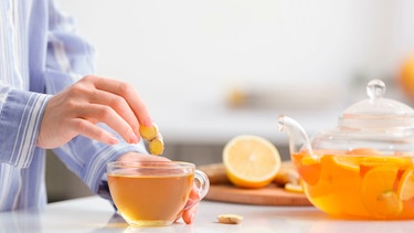 Die Hand einer Frau hält eine Scheibe Ingwer über eine Tasse Ingwertee, im Hintergrund die Teekanne und eine halbe Zitrone auf einem Schneidebrett | Bild: mauritius Bilder / Pixel-shot / Alamy / Alamy Stock Fotos