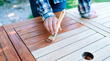 Hand mit einem Pinsel streicht einen Gartentisch | Bild: mauritius images / Wojciech Kozielczyk / Alamy / Alamy Stock Photos