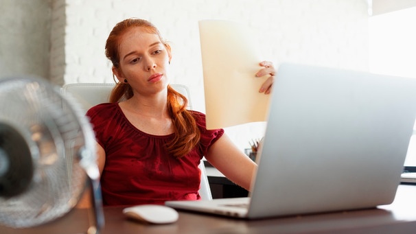 Eine Frau sitzt während einer Hitzewelle im Büro und fächert sich Luft zu. | Bild: mauritius images / DCPhoto / Alamy / Alamy Stock Photos