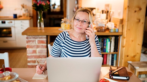 Frau sitzt mit ihrem Laptop und Handy am Ohr zu Hause im Homeoffice | Bild: mauritius-images