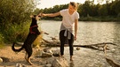 Eine Frau steht barfuss im Fluss und spielt mit ihrem Hund. | Bild: mauritius images / Westend61 / Christina Falkenberg