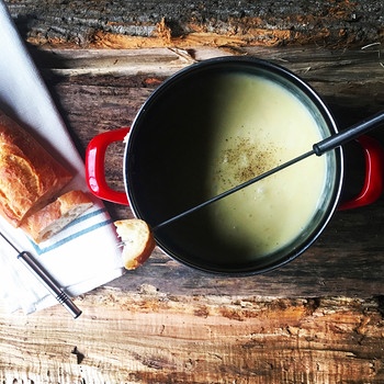 Auf hölzernem Untergrund steht ein Topf mit Käsefondue. Daneben liegt ein angebrochenes Baguette. | Bild: mauritius images / Cavan Images